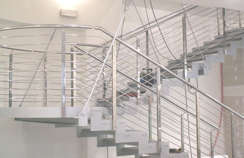 Schody wewnętrzne z balustradą ze stali nierdzewnej, widoczne elementy montażowe i przewody elektryczne sugerujące etap prac wykończeniowych.